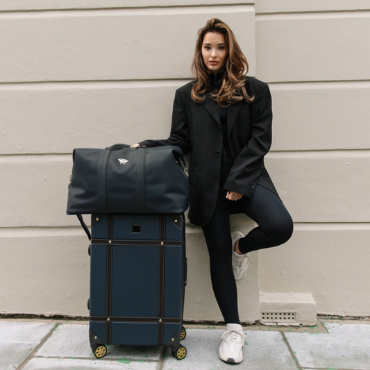 Woman in black coat with navy blue suitcase and duffel bag against a beige wall.